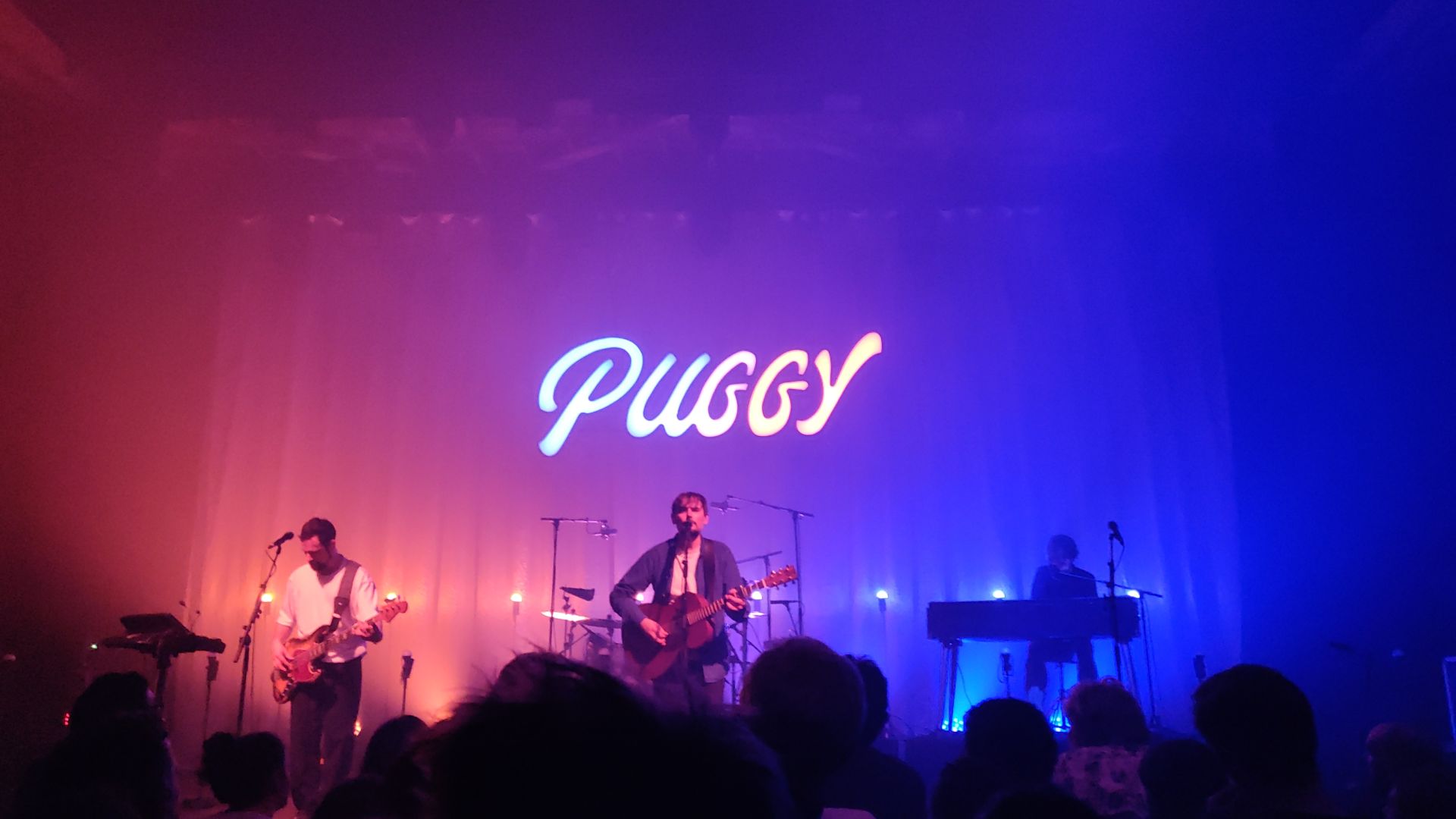 trois musiciens sur scène avec écrit Puggy en lettres lumineuses.